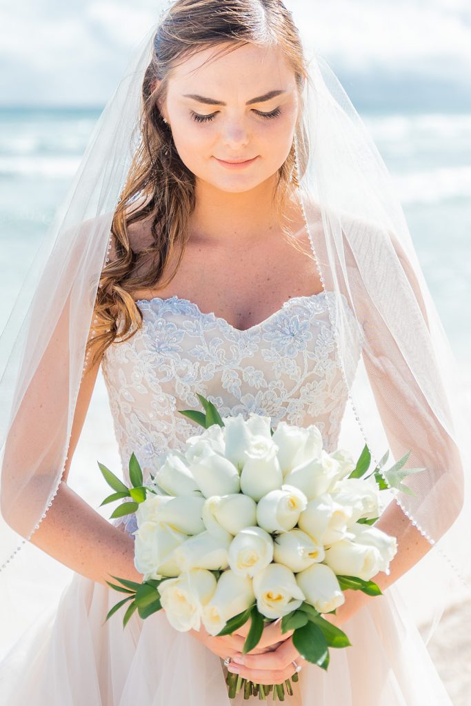 a portrait image of a bride on the beach wearing an off-white wedding dress with a princess cut neckline.  She is holding a bouquet of white roses.