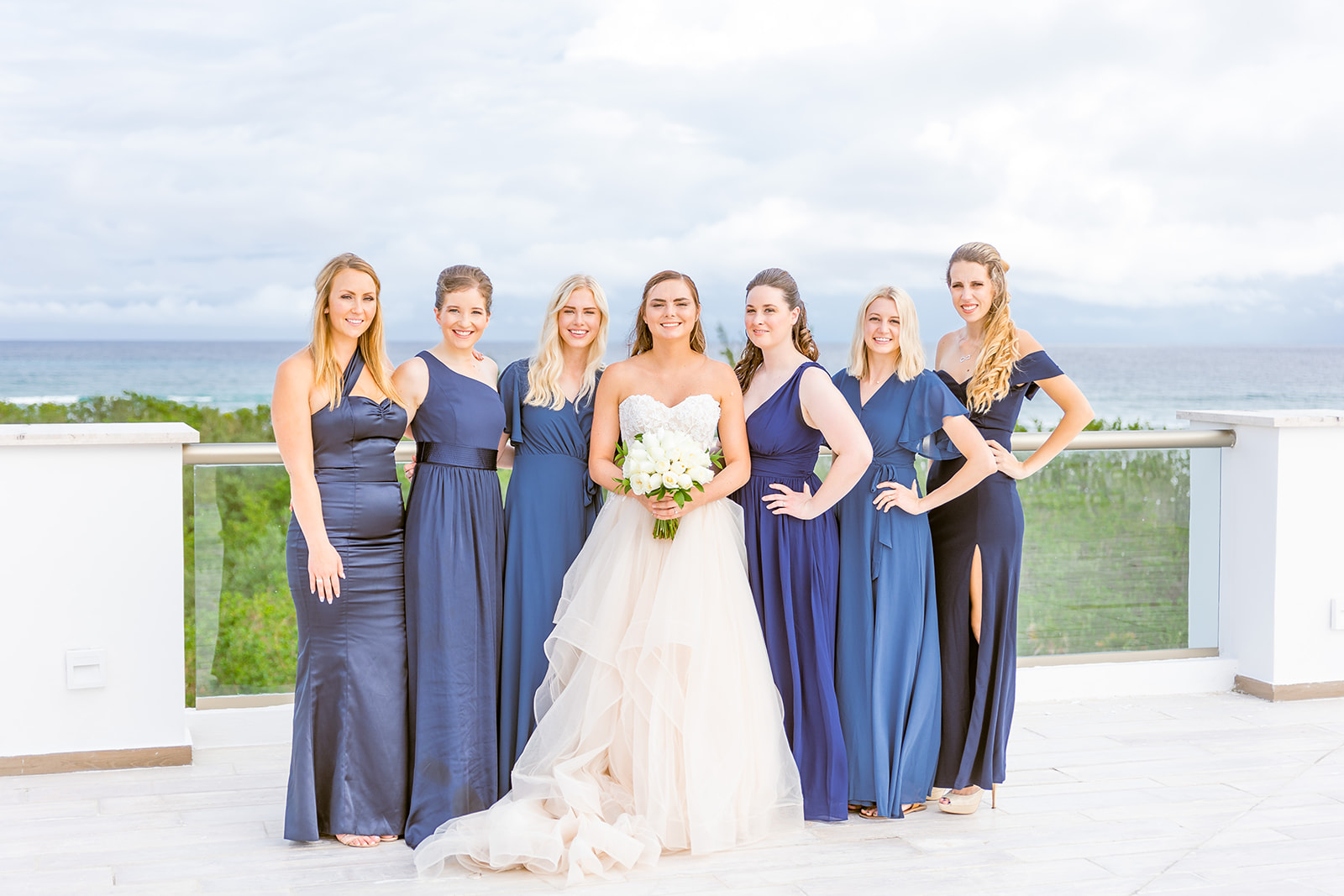 A portrait image of a bride and her bridesmaids posing for the camera in front of a beach in Yucatan, Mexico.   The bride is wearing an off-white, princess cut A-line wedding dress and holding a bouquet of white roses.  The bridesmaids are all wearing long navy blue dresses.