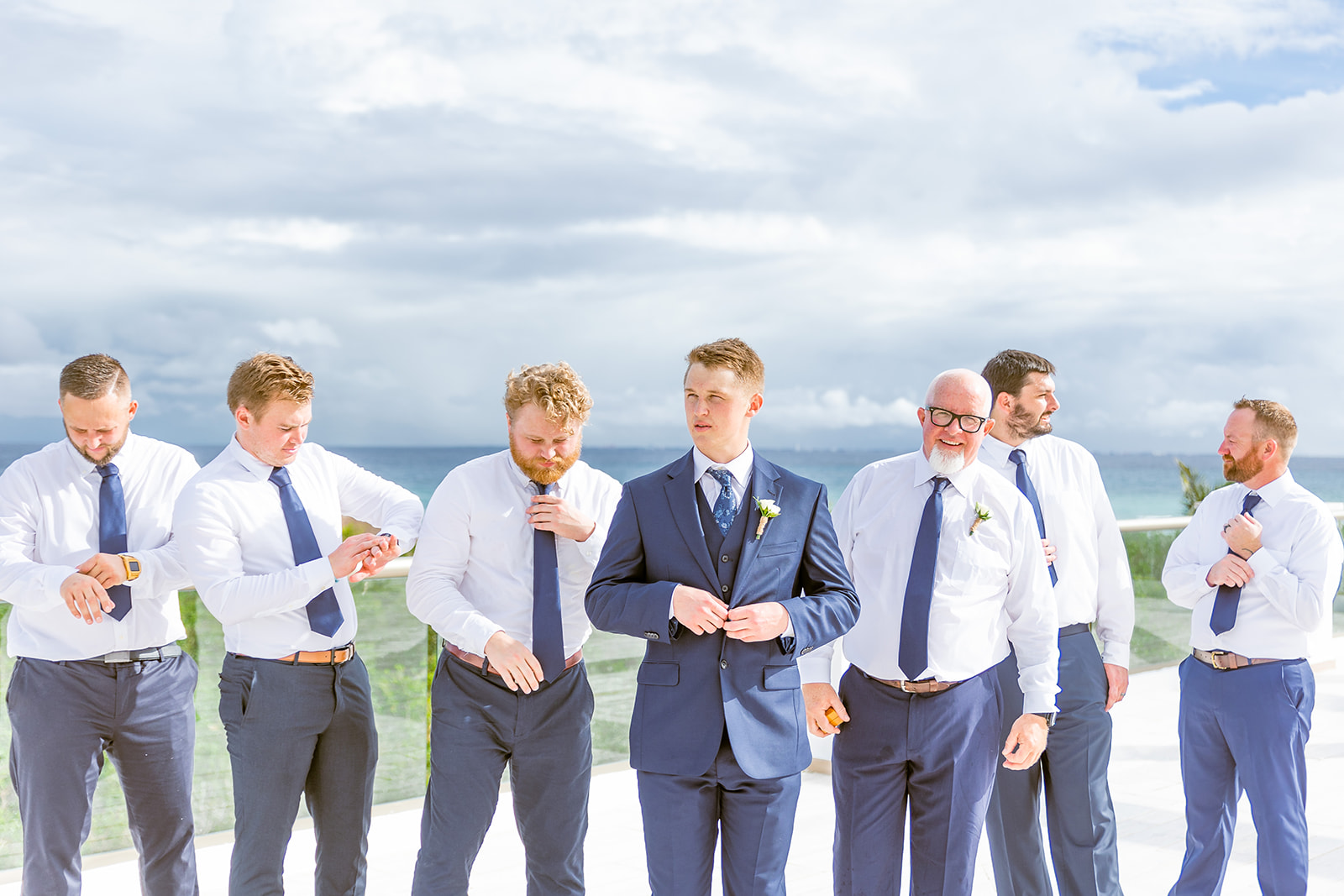 A portrait of a groom and his groomsmen.  The groom is wearing a navy blue suit and the groomsmen are wearing navy blue pants and tie with white shirts and no jackets.